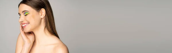 Pretty teen girl with multicolored visage and naked shoulders touching face isolated on grey, banner — Stock Photo