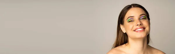 Teen girl with naked shoulders and multicolored makeup looking at camera isolated on grey, banner — Stock Photo