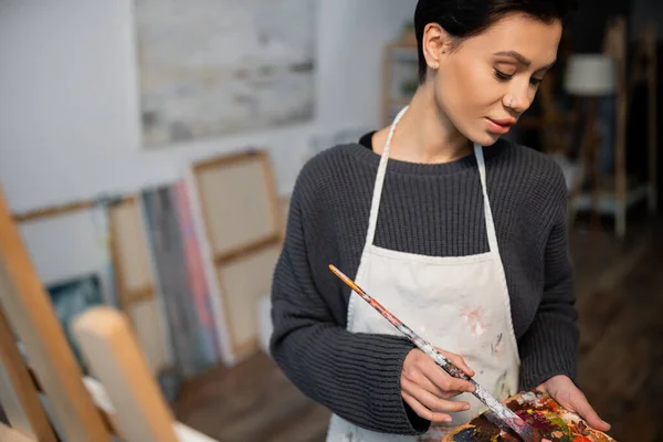 Brunette artist holding paintbrush and palette near blurred easel - foto de stock