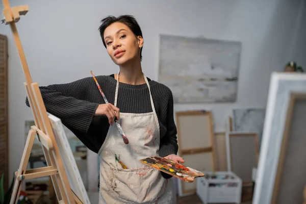 Artist in apron holding palette and paintbrush while looking at camera in workshop — Foto stock