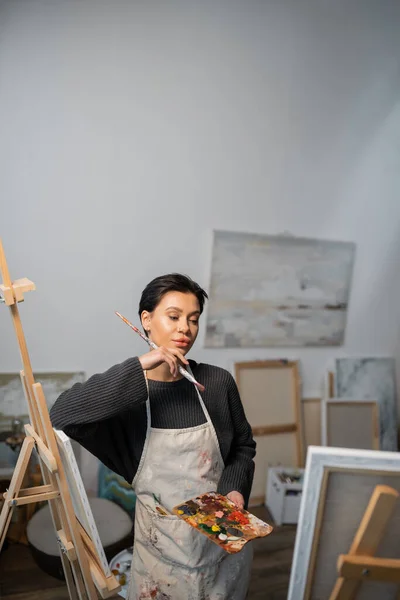 Focused artist holding palette and looking at painting in workshop — Stock Photo