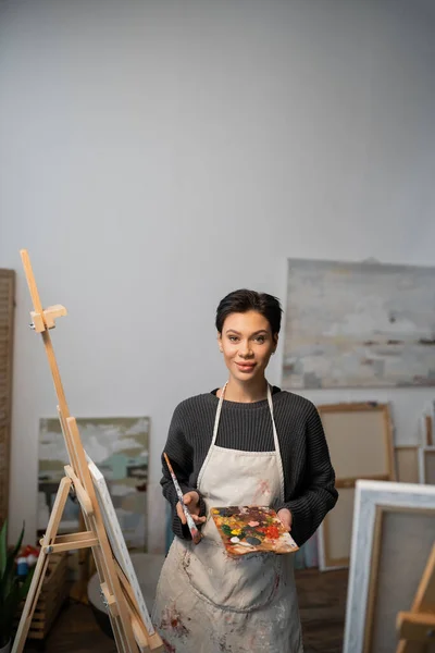 Young artist holding palette with paints and looking at camera near easels in workshop — Stock Photo