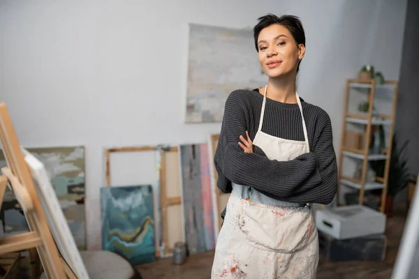 Artist in apron crossing arms and looking at camera in workshop — Stock Photo
