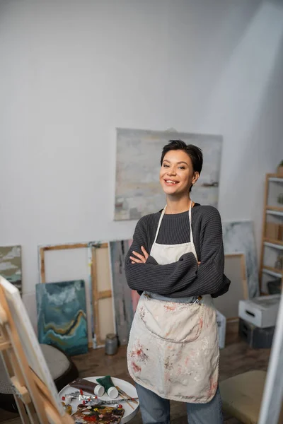 Cheerful artist in apron looking at camera near paints and easel in studio — Stock Photo