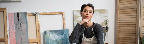 Brunette artist looking at camera near paintings in studio, banner — Stock Photo