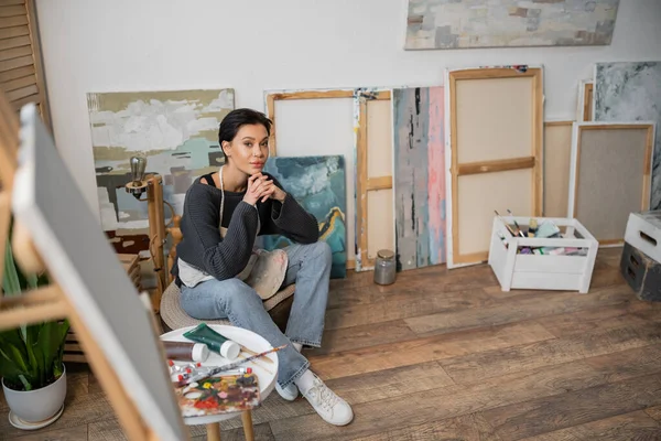 Молодой художник смотрит в камеру, сидя рядом с красками в мастерской — стоковое фото
