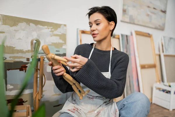 Artist in apron holding wooden doll near blurred paintings in studio - foto de stock