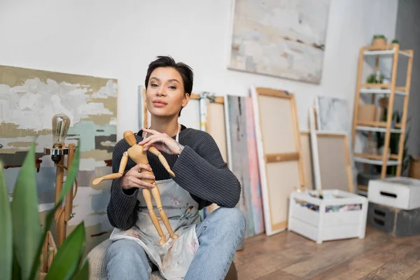 Молодой художник держит деревянную куклу и смотрит в камеру рядом с картинами в студии — стоковое фото
