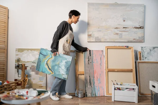 Вид на молодого художника в фартуке, смотрящего на картины в мастерской — стоковое фото