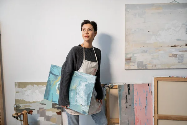 Dreamy artist in apron looking away near paintings in studio - foto de stock