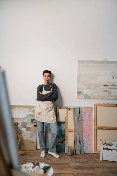 Young artist in apron crossing arms while standing near paintings in studio — Foto stock