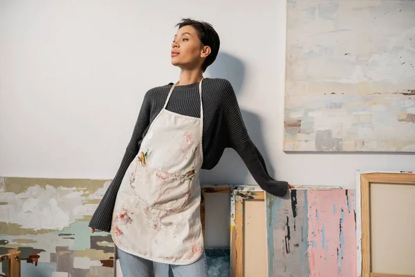 Brunette artist in apron looking away near paintings in studio — Stock Photo