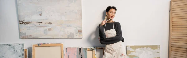 Dreamy young artist holding paintbrushes near drawings in workshop, banner — Stock Photo