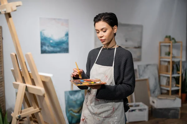 Young artist in apron holding paintbrush and palette near canvas on easel — Stock Photo