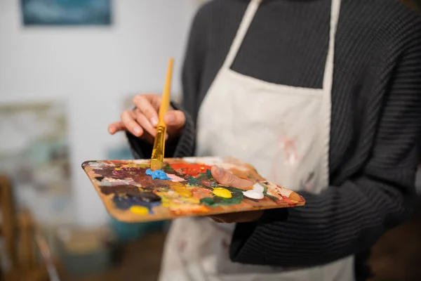 Cropped view of blurred artist in apron holding palette and paintbrush in studio — Stock Photo
