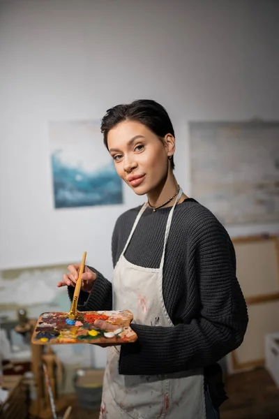 Portrait of short haired artist in apron holding paintbrush and palette in workshop — Stock Photo