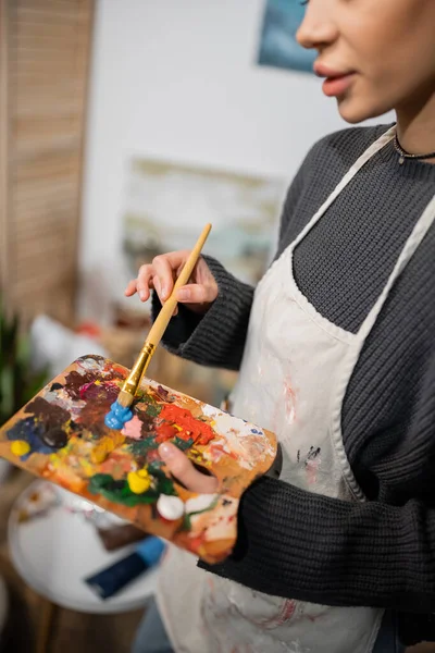 Cropped view of artist in apron mixing paints on palette in workshop — Stock Photo