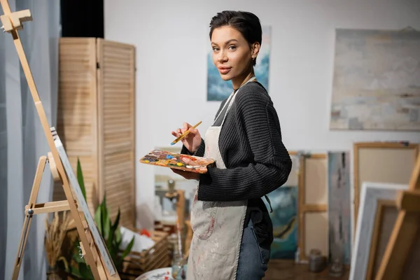 Young artist looking at camera while holding palette near easel in workshop - foto de stock