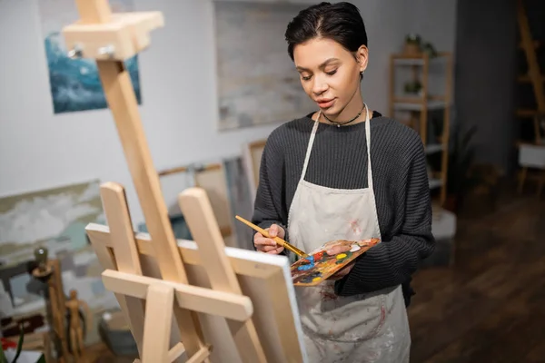 Short haired artist mixing paints on palette near blurred easel in workshop — Stock Photo
