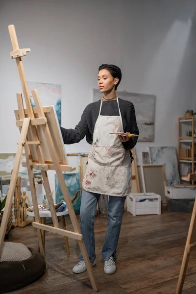 Artist in apron holding palette and looking at canvas on easel in studio — Stock Photo