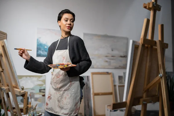 Short haired artist in apron holding paintbrush and palette near canvases on easels in studio - foto de stock
