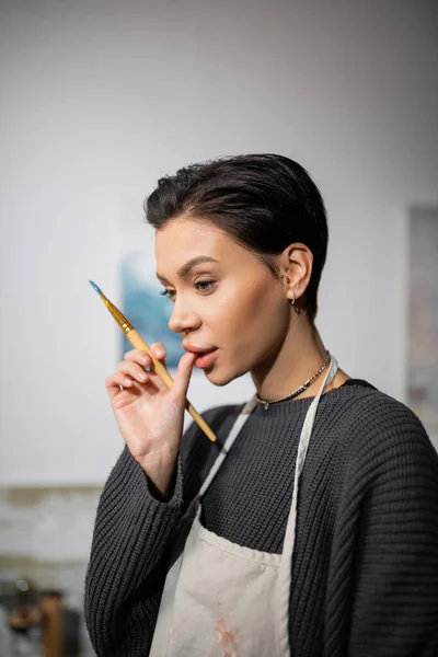 Thoughtful artist holding paintbrush and touching lips in workshop — Stock Photo