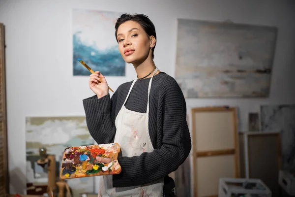 Brunette artist looking at camera while holding paintbrush and palette in blurred workshop — Stock Photo