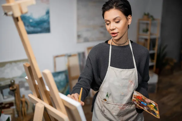 Short haired artist in apron holding palette while painting on blurred canvas — Fotografia de Stock