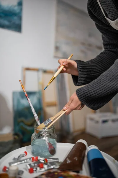 Cropped view of artist taking paintbrushes from jar with water in workshop — Photo de stock