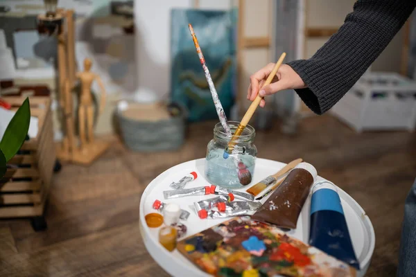Cropped view of artist taking paintbrush from jar with water near paints in studio — Stock Photo