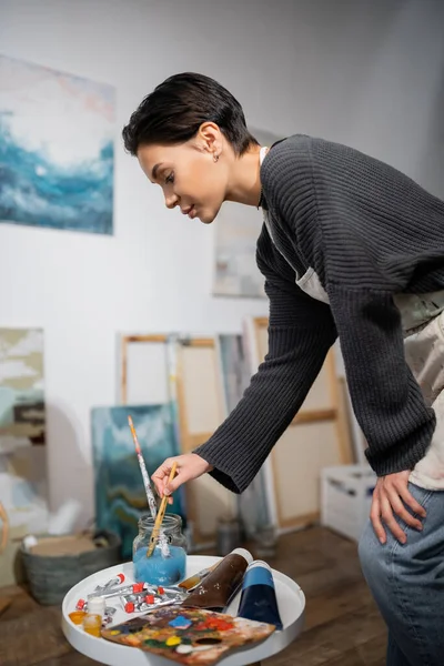 Side view of young artist taking paintbrush from jar near palette and paints in studio — Stockfoto
