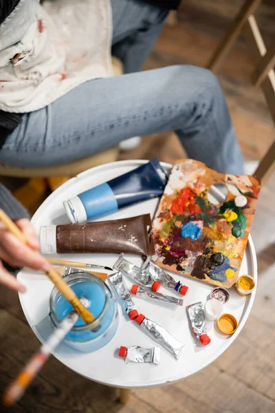 Вид зверху художника в фартух покласти пензлик в банку з водою біля фарб на столі в майстерні — стокове фото