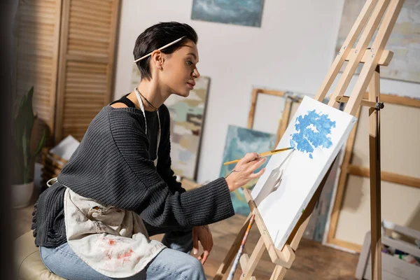 Side view of young artist in apron painting on canvas — Stock Photo