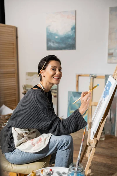Positive artist looking at camera while holding paintbrush near canvas in studio — Stock Photo