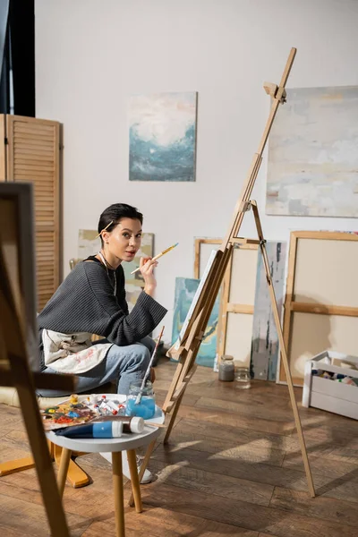 Brunette artist holding paintbrush and looking at camera near canvas on easel in studio — Stock Photo