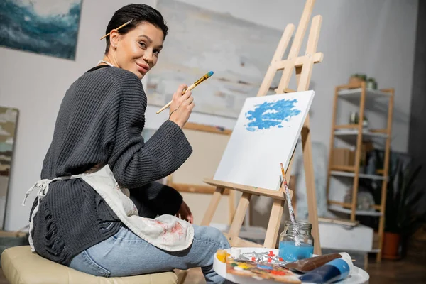 Pleased artist holding paintbrush and looking at camera near easel on canvas — Stock Photo