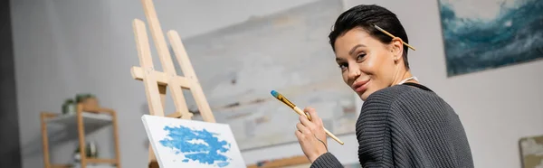 Positive artist with paintbrushes looking at camera near canvas, banner — Stock Photo