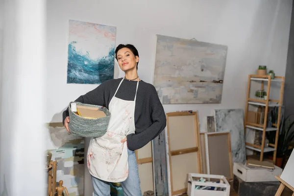 Short haired artist in apron holding basket with equipment near drawings in studio — Stock Photo