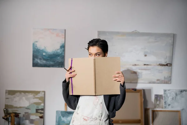 Brunette artist in dirty apron holding sketchbook near face in studio — Stock Photo