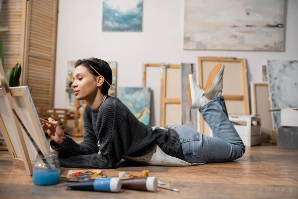 Young artist painting on canvas while lying near pains and palette on floor in workshop — Stock Photo