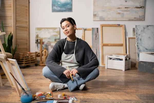 Young artist in apron holding paintbrush near paints and canvas in workshop — Stock Photo