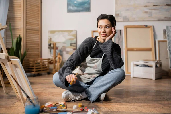 Positive artist in apron holding paintbrush near canvas and easel in workshop — Foto stock