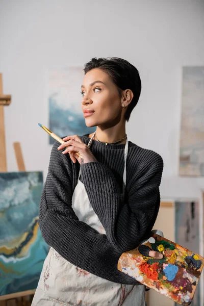 Short haired artist in sweater and apron holding palette with paints and paintbrush - foto de stock
