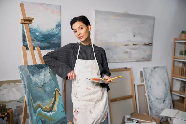 Brunette artist holding paintbrush and palette near canvas on easel in studio — Foto stock