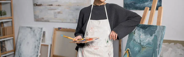 Cropped view of artist in apron holding palette near painting on easel in studio, banner — Foto stock