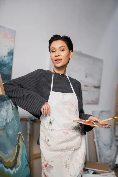 Brunette artist in dirty apron holding palette near drawing in workshop — Stock Photo