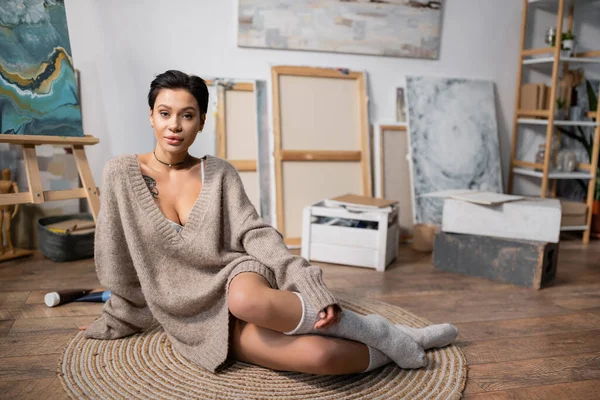 Sexy artist in sweater looking at camera while sitting on floor in studio — Stockfoto