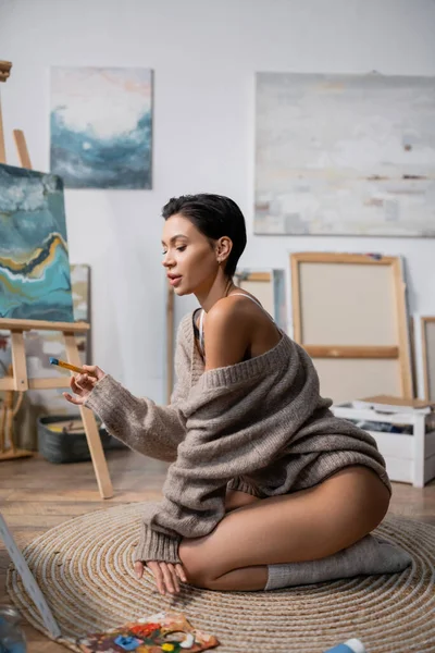 Sexy artist holding paintbrush near palette on floor in studio — Stock Photo