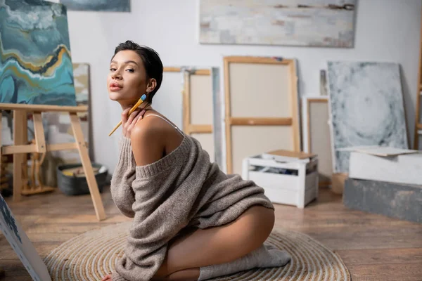 Seductive artist in sweater holding paintbrush in workshop — Stockfoto
