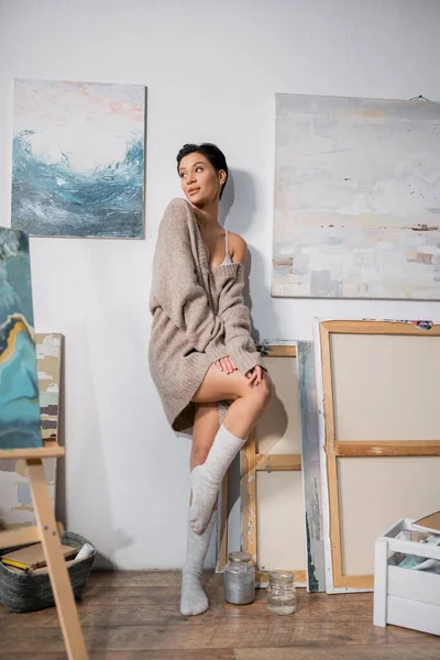 Seductive artist in socks and sweater looking away near drawings in workshop — Stock Photo